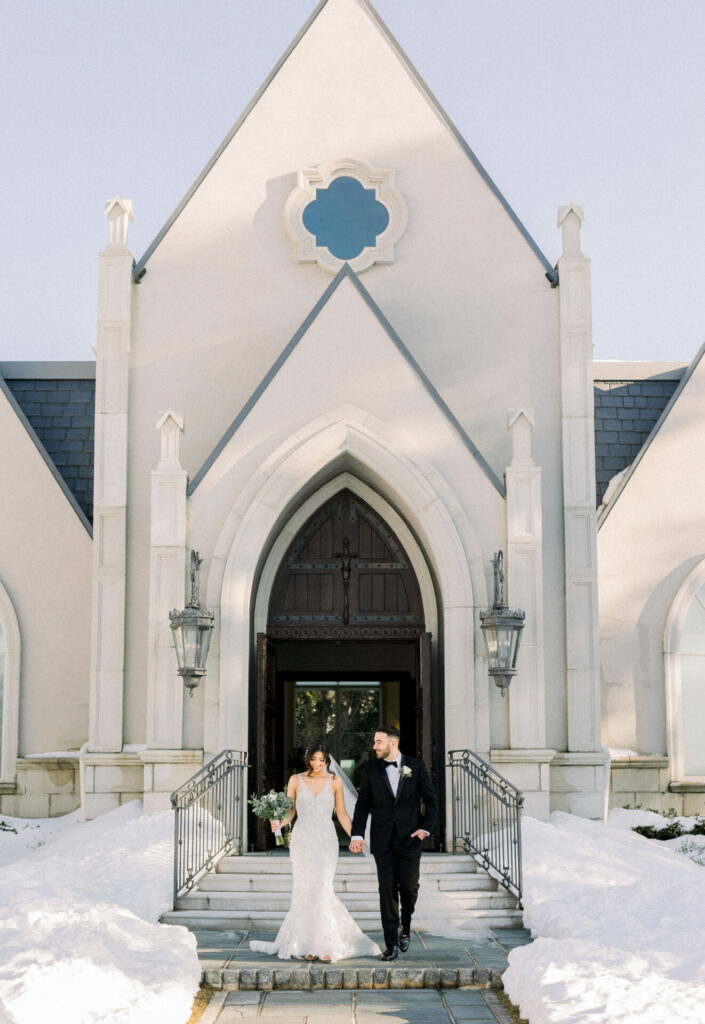 Bride and groom holding hands, smiling on snowy church steps.