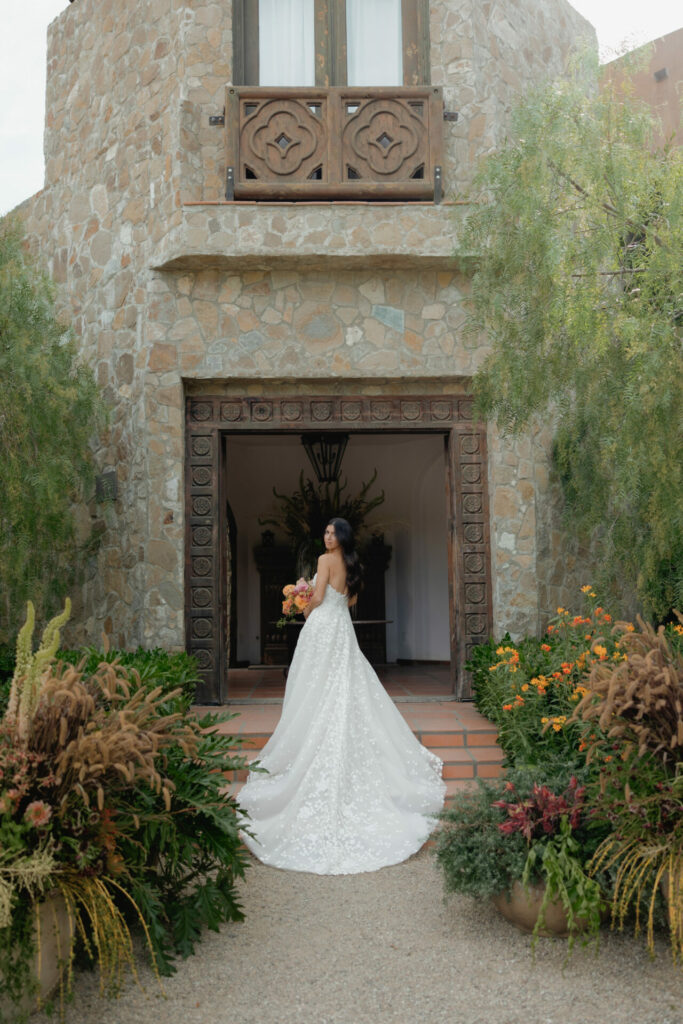 Bride in floral wedding gown by rustic stone building.