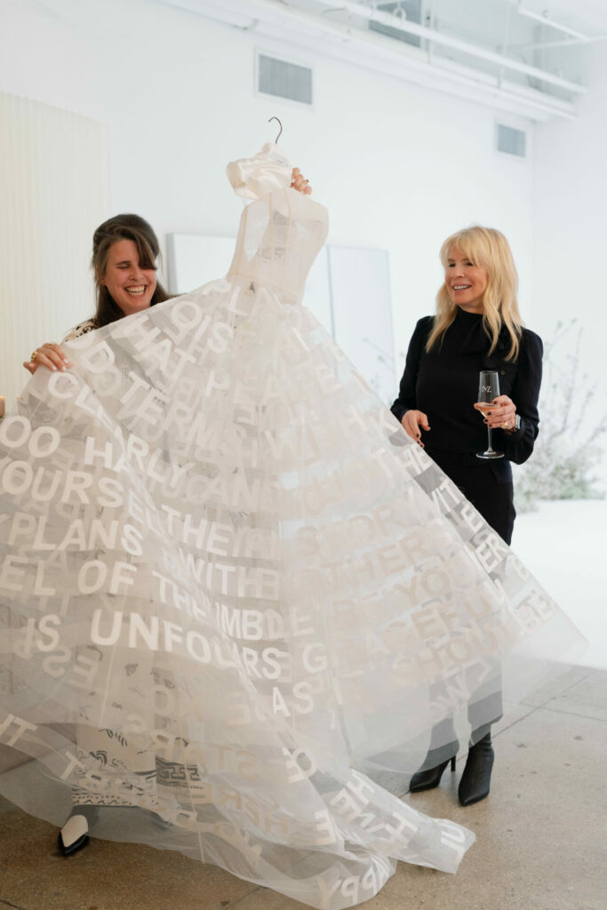 Two women smile; one shows dress, one holds wine.