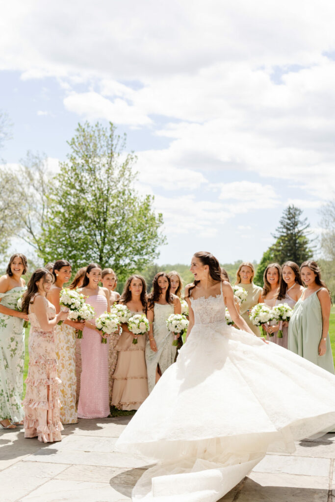 Bride twirls outdoors, bridesmaids in pastel dresses nearby.