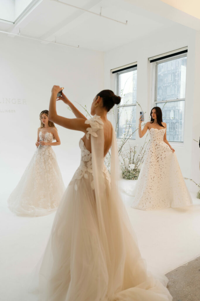 Three women in white bridal gowns pose; one takes a selfie.