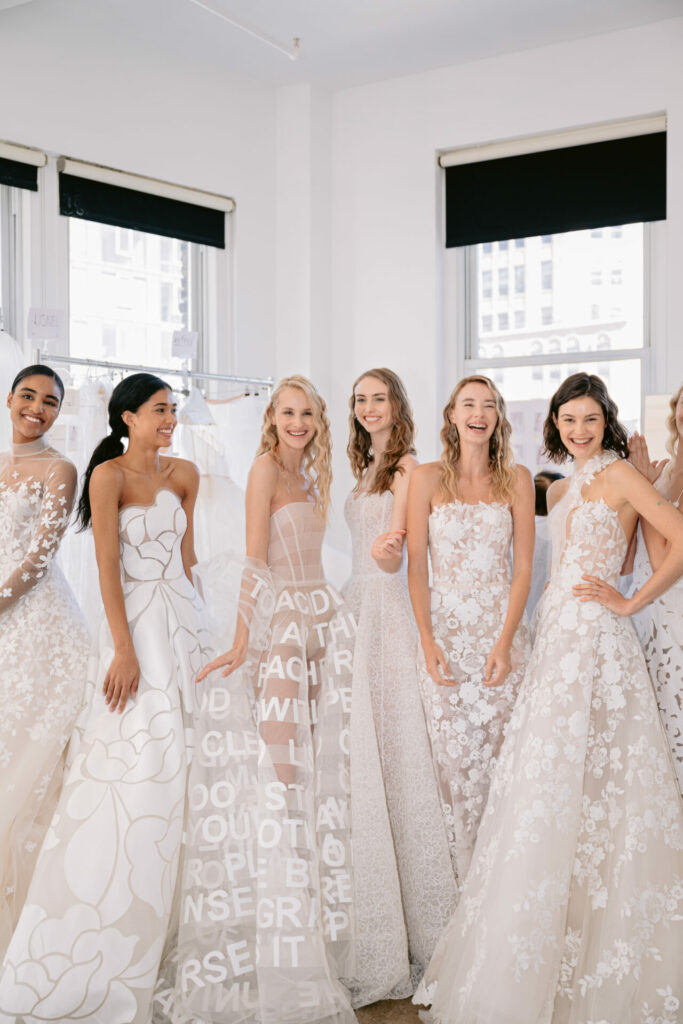 Six women in patterned wedding gowns laugh together in sunlight.
