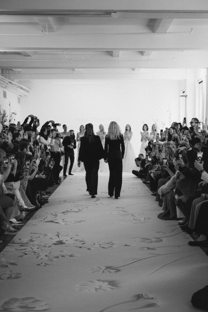 Two women walk hand-in-hand down floral runway.