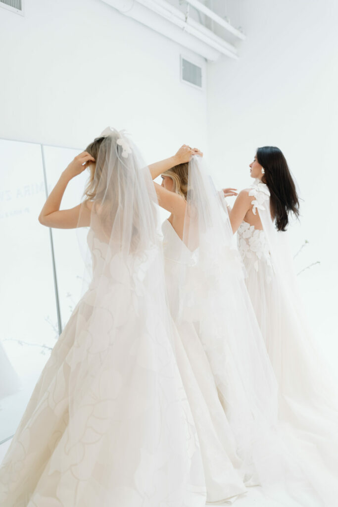 Three women in floral bridal gowns face mirror.