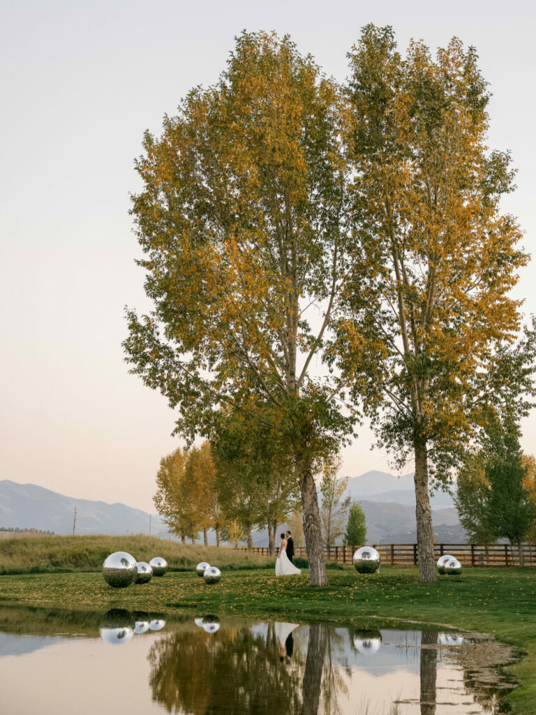 Bride and groom under autumn trees by pond, sculptures nearby.
