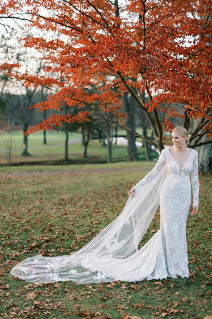 Bride in lace dress under orange autumn tree outdoors.