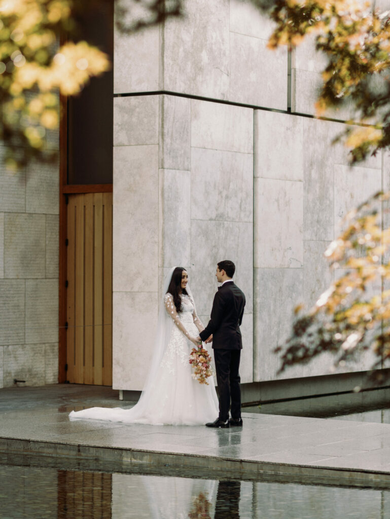 Bride and groom hold hands by modern stone building.