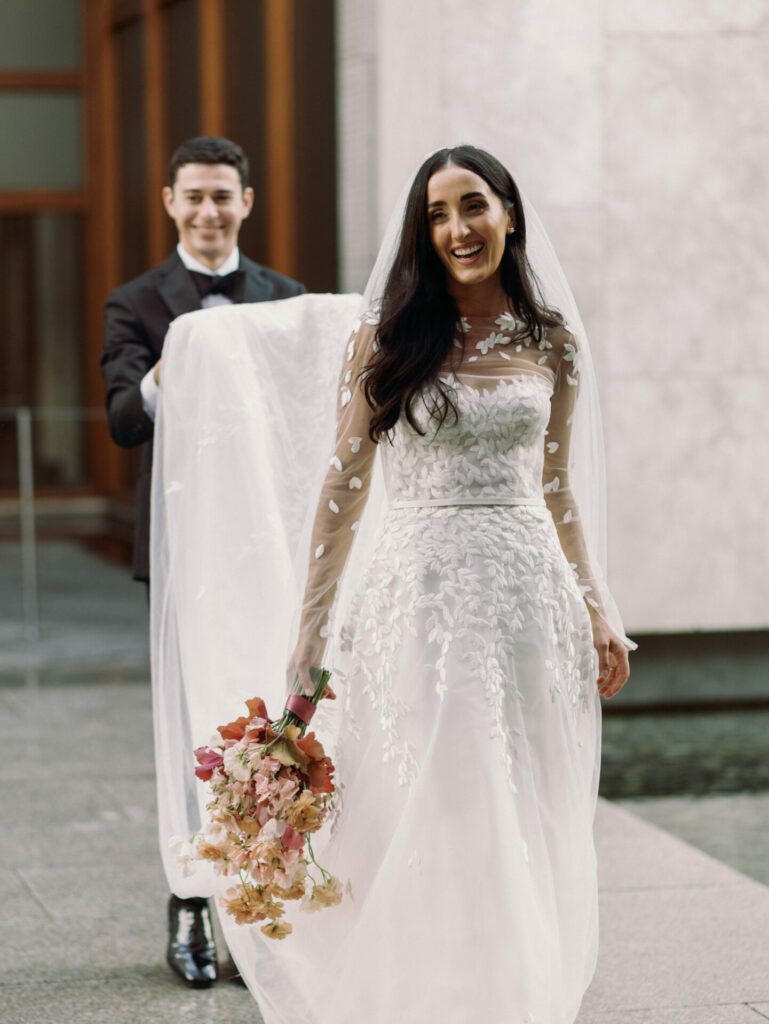 Bride in embroidered gown, veil carried outdoors.