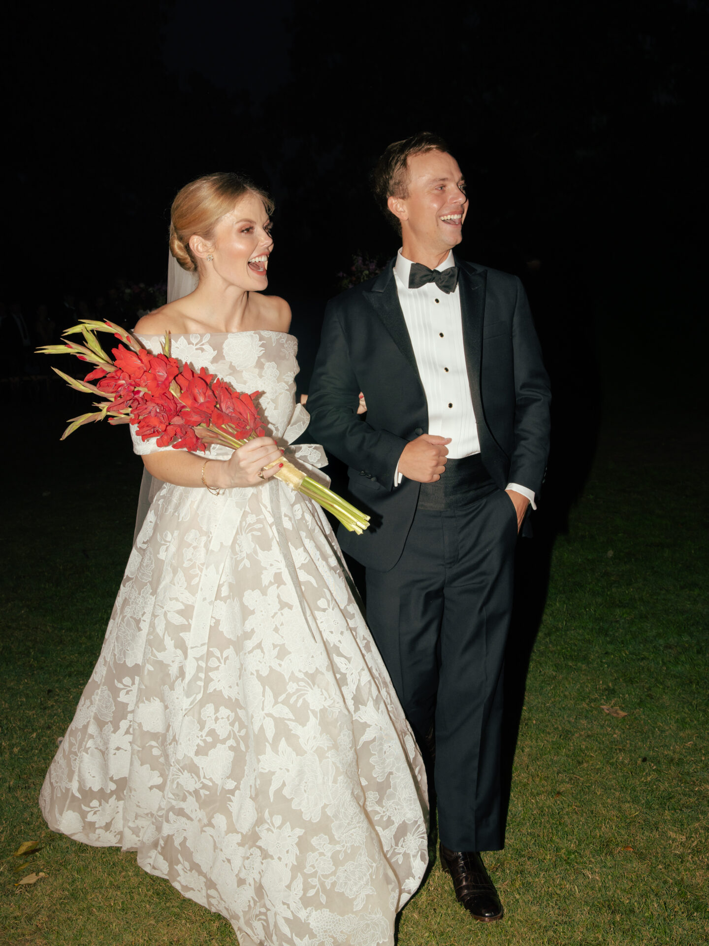 Bride in floral gown laughs with groom at night.