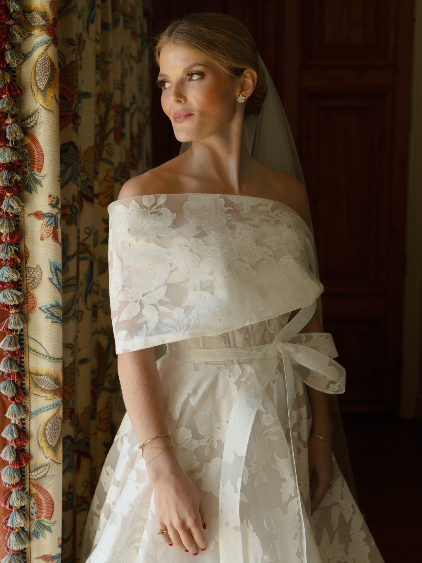 Bride in lace dress gazes out sunlit window.