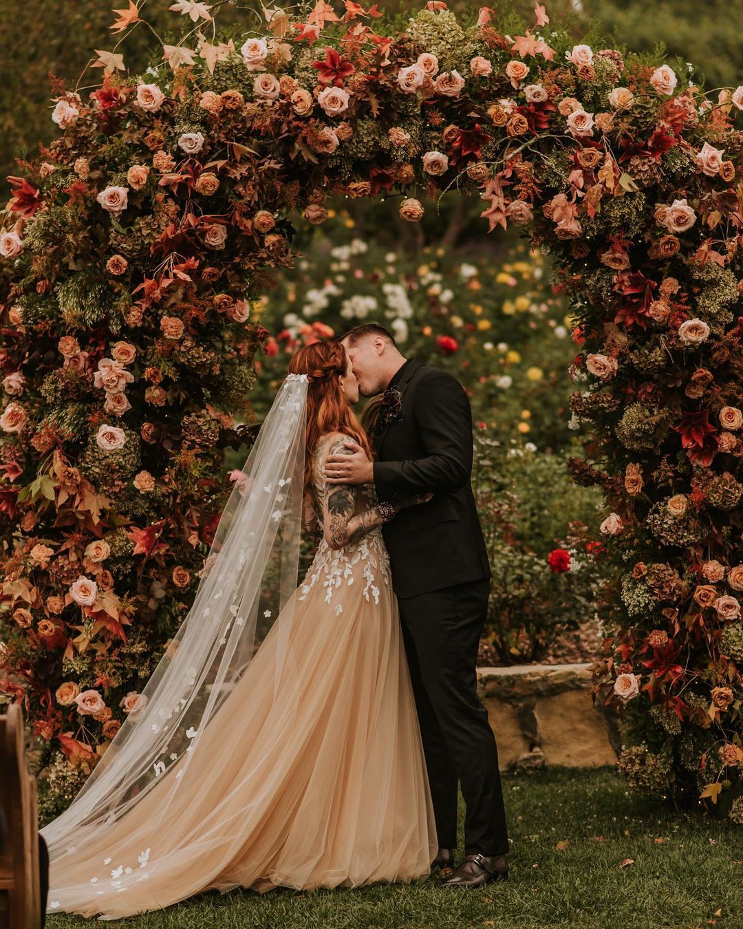 Bride and groom kiss beneath autumn floral arch.