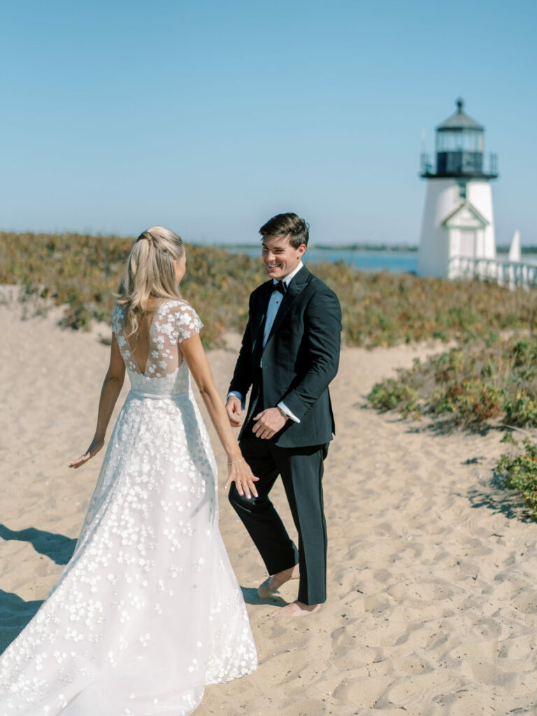 Bride and groom smiling on beach by lighthouse.