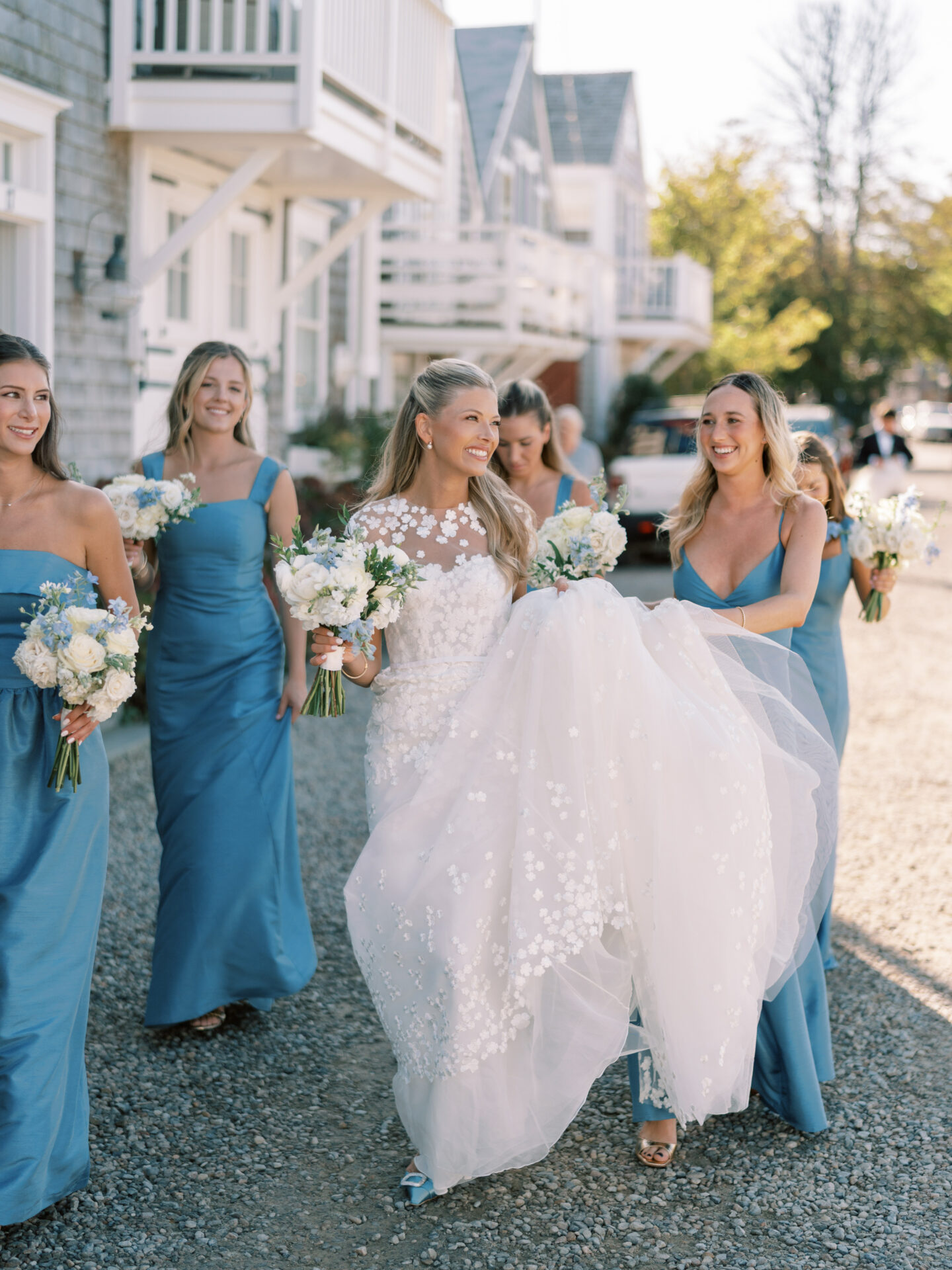 Bride in floral gown with blue-dressed bridesmaids smiling outdoors.