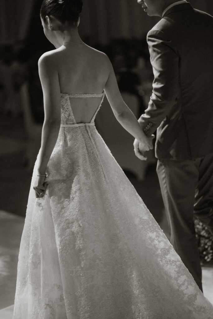 Bride and groom walk hand-in-hand, seen from behind.