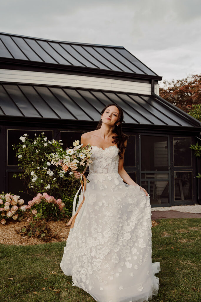 Bride in lace gown, smiling with bouquet by house.