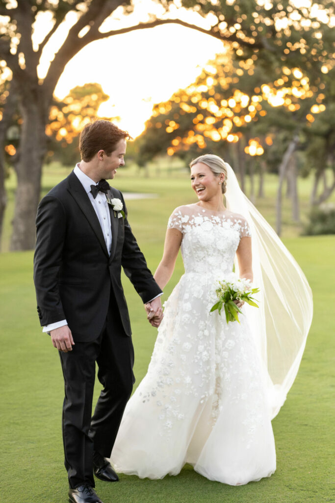 Bride and groom hold hands, walk on sunset lawn.