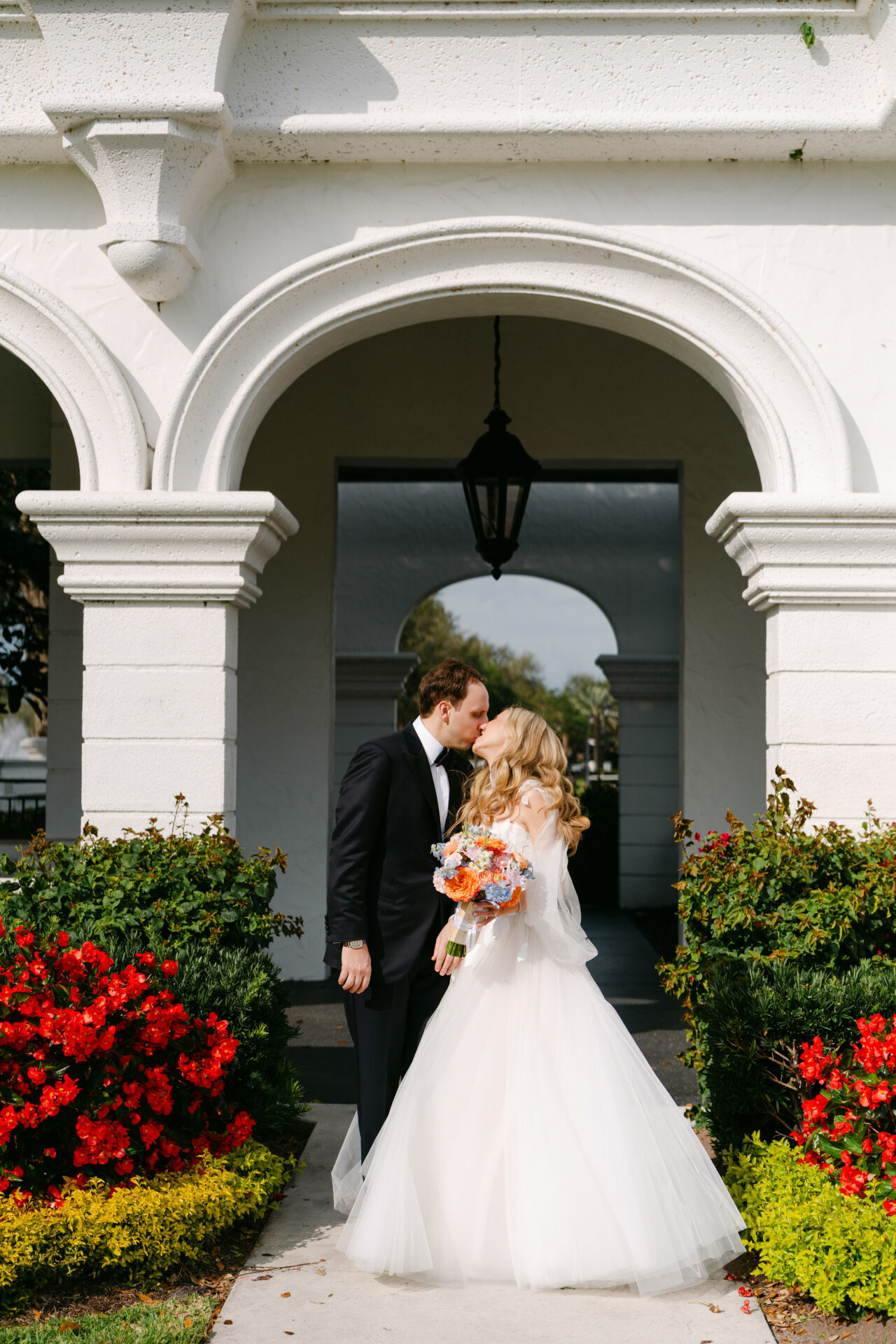 Bride and groom kiss by white arch, flowers bloom.