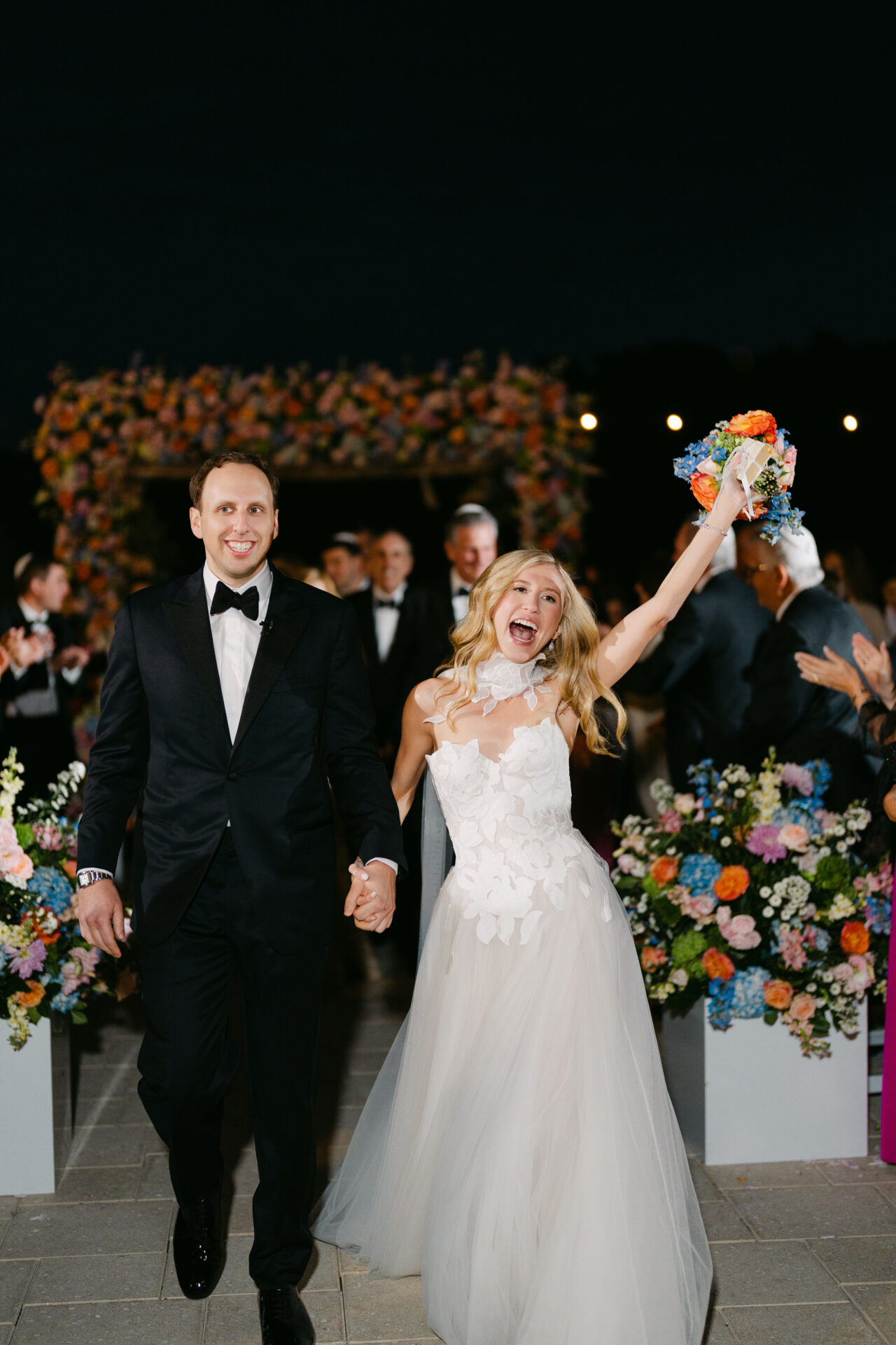 Smiling bride and groom walk down aisle at night.