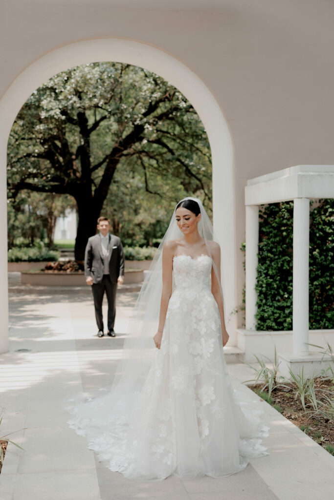 Bride in couture gown smiles under archway; groom waits.