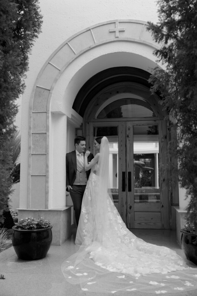 Bride in Mira Zwillinger gown smiles with groom by arched doorway.