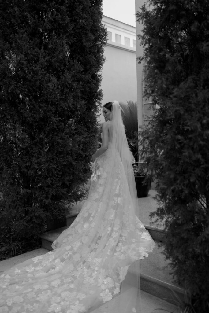 Bride in lace gown, veil, poses outdoors on steps.