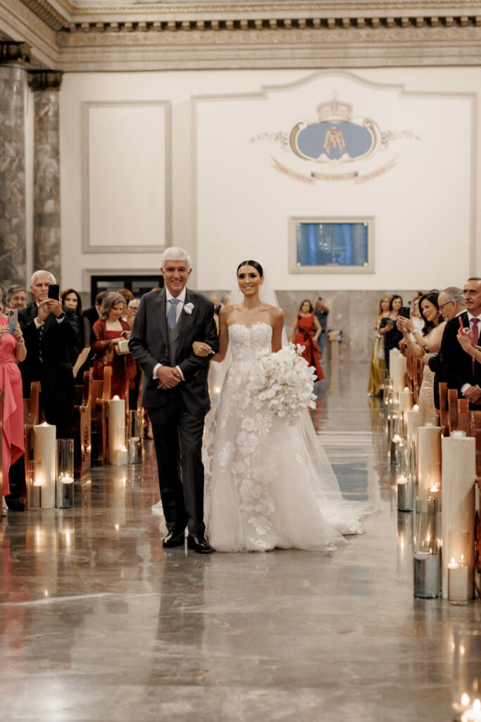 Bride in designer gown walks aisle, guests watch.