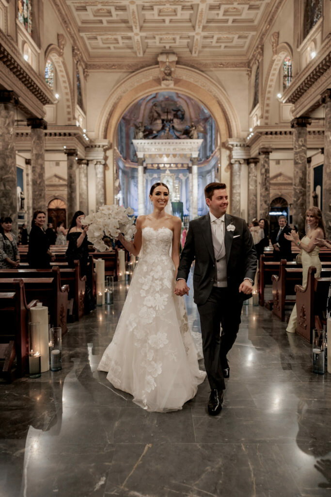 Smiling newlyweds walk the aisle as guests applaud.