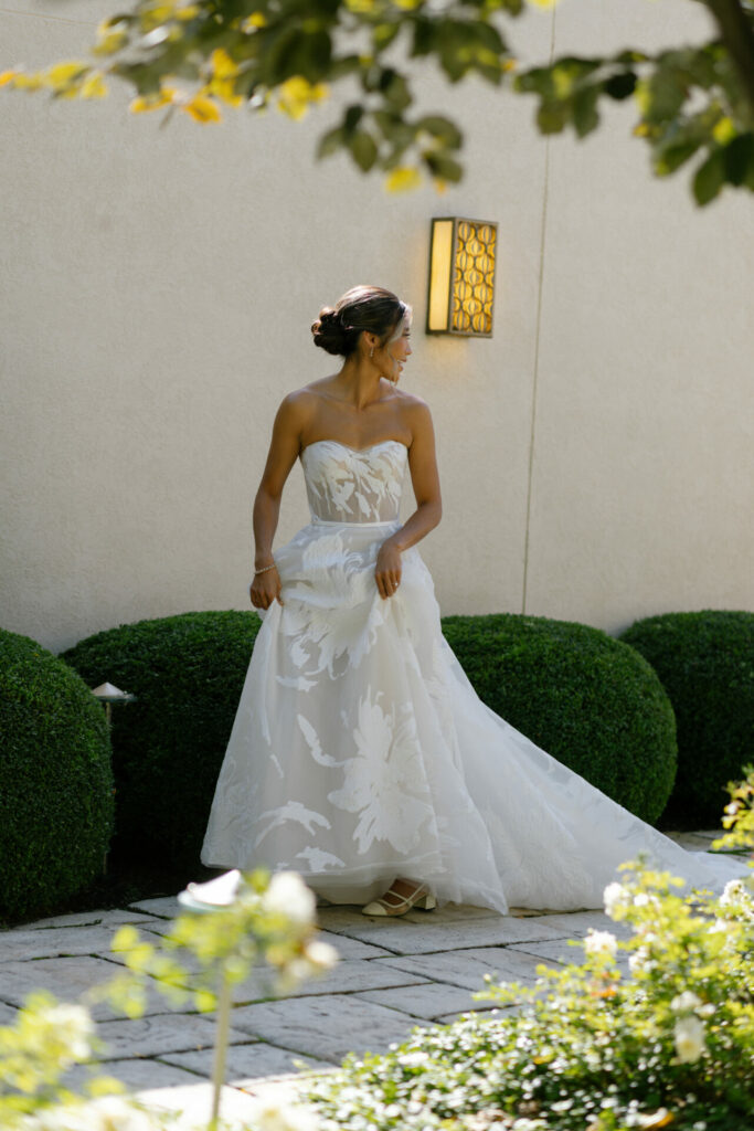 Bride in white gown stands on garden path.