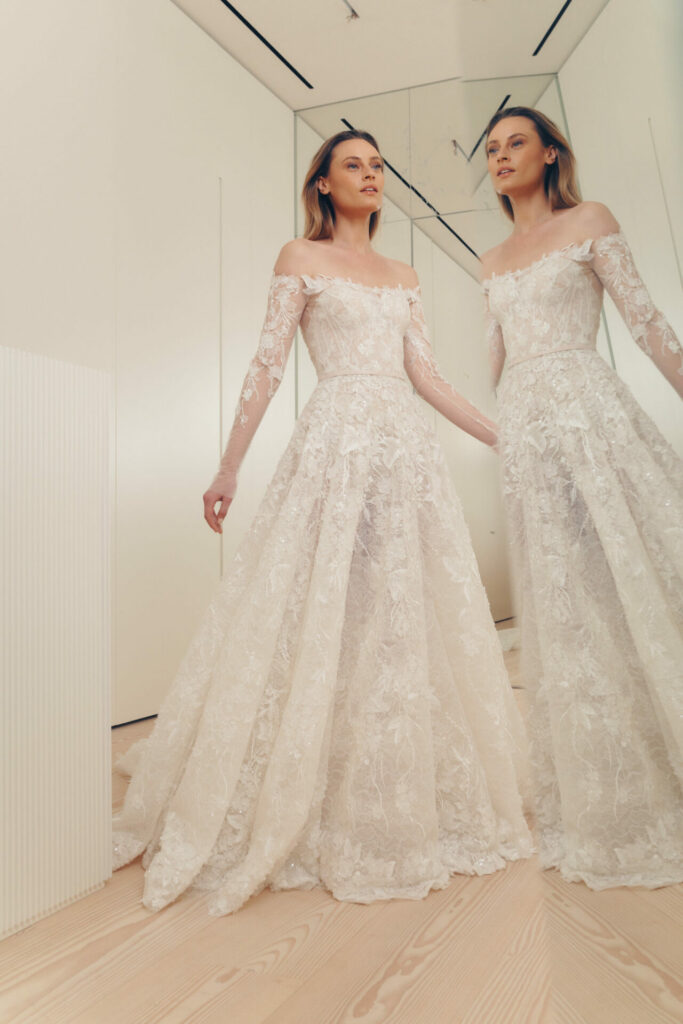 Bride in lace gown stands by mirror, reflected.