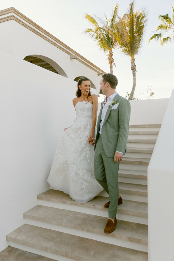 Bride and groom holding hands, smiling on outdoor stairs.