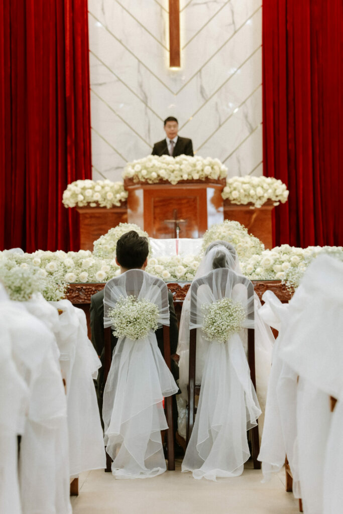 Bride and groom seated before altar of white flowers.