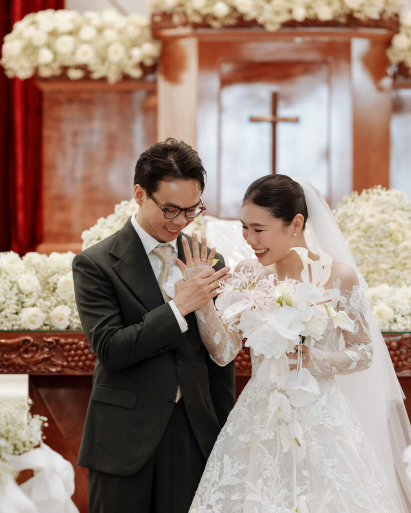 Bride and groom smile, admiring ring amid white flowers.