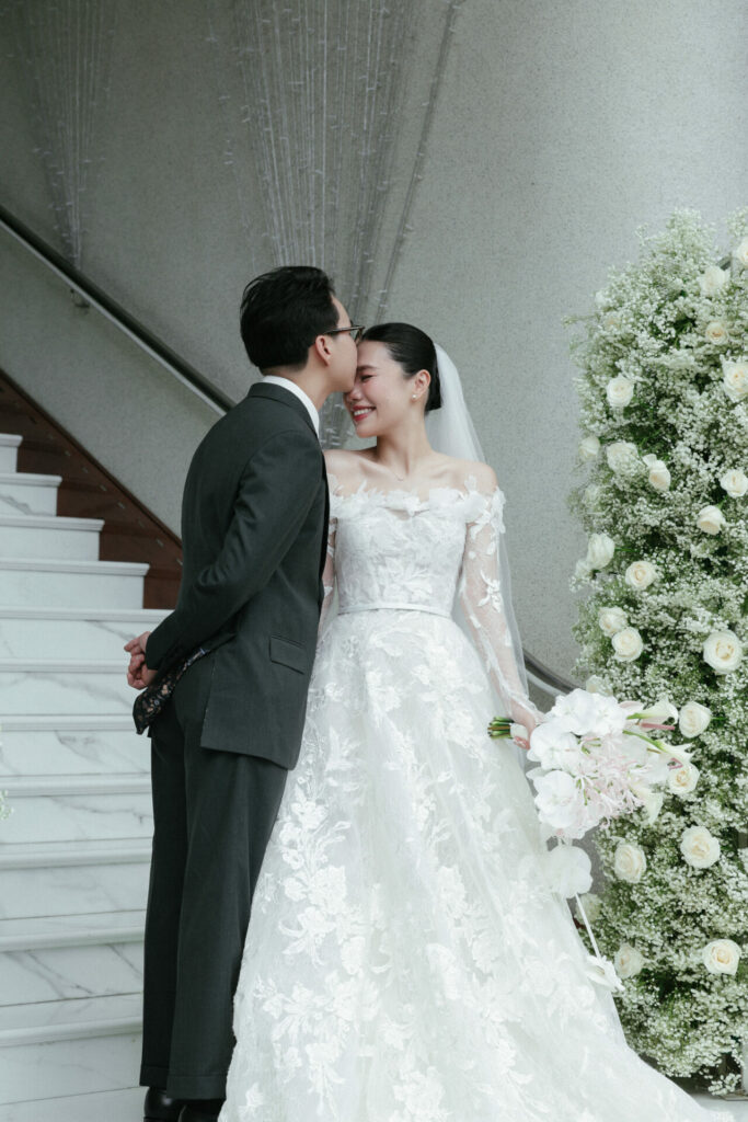 Katya Ruiz in Mira Zwillinger gown, smiling on floral staircase.