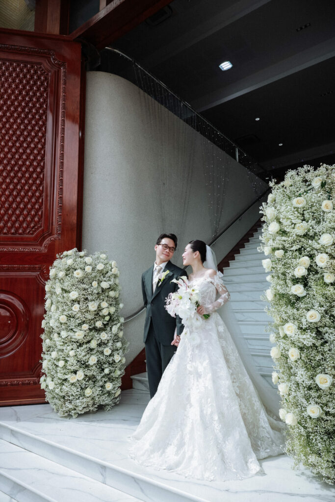 Bride and groom on marble steps amid white flowers.