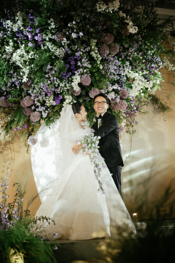 Katya Ruiz and groom embrace under purple, white flowers.