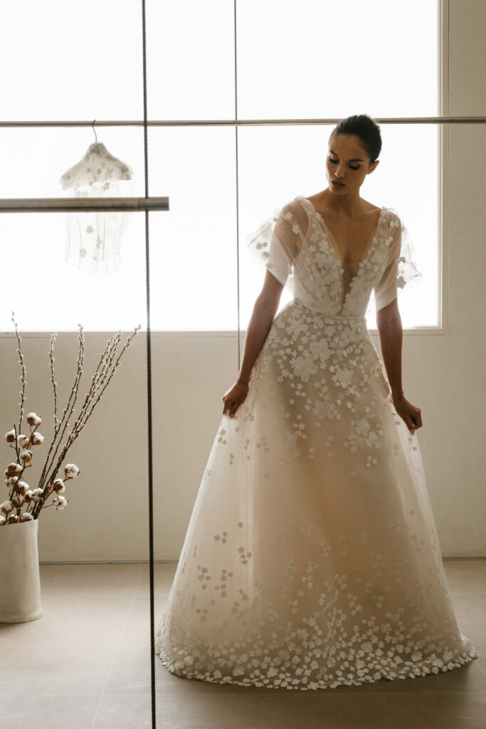 Bride in floral dress near window, cape behind.