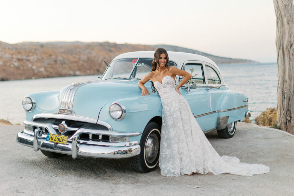 Bride in white gown by blue car, seaside hills.