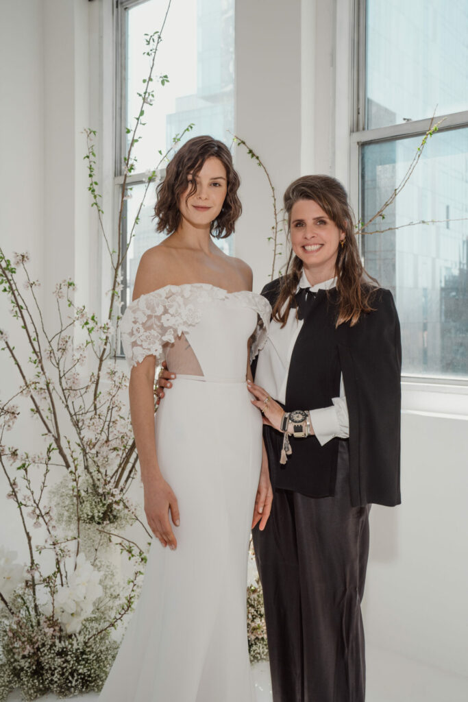 Smiling Ness & Nessia in wedding outfits by windows.