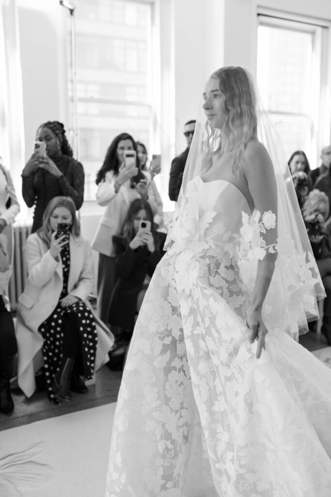Bride in lace gown walks runway as crowd films.