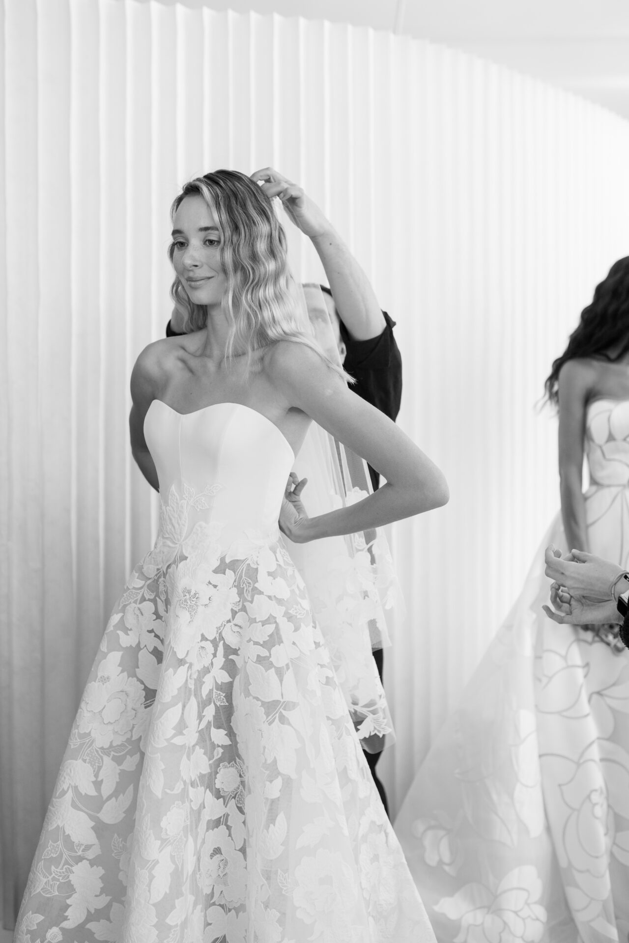 Two women in floral strapless bridal gowns, one hairstyling.
