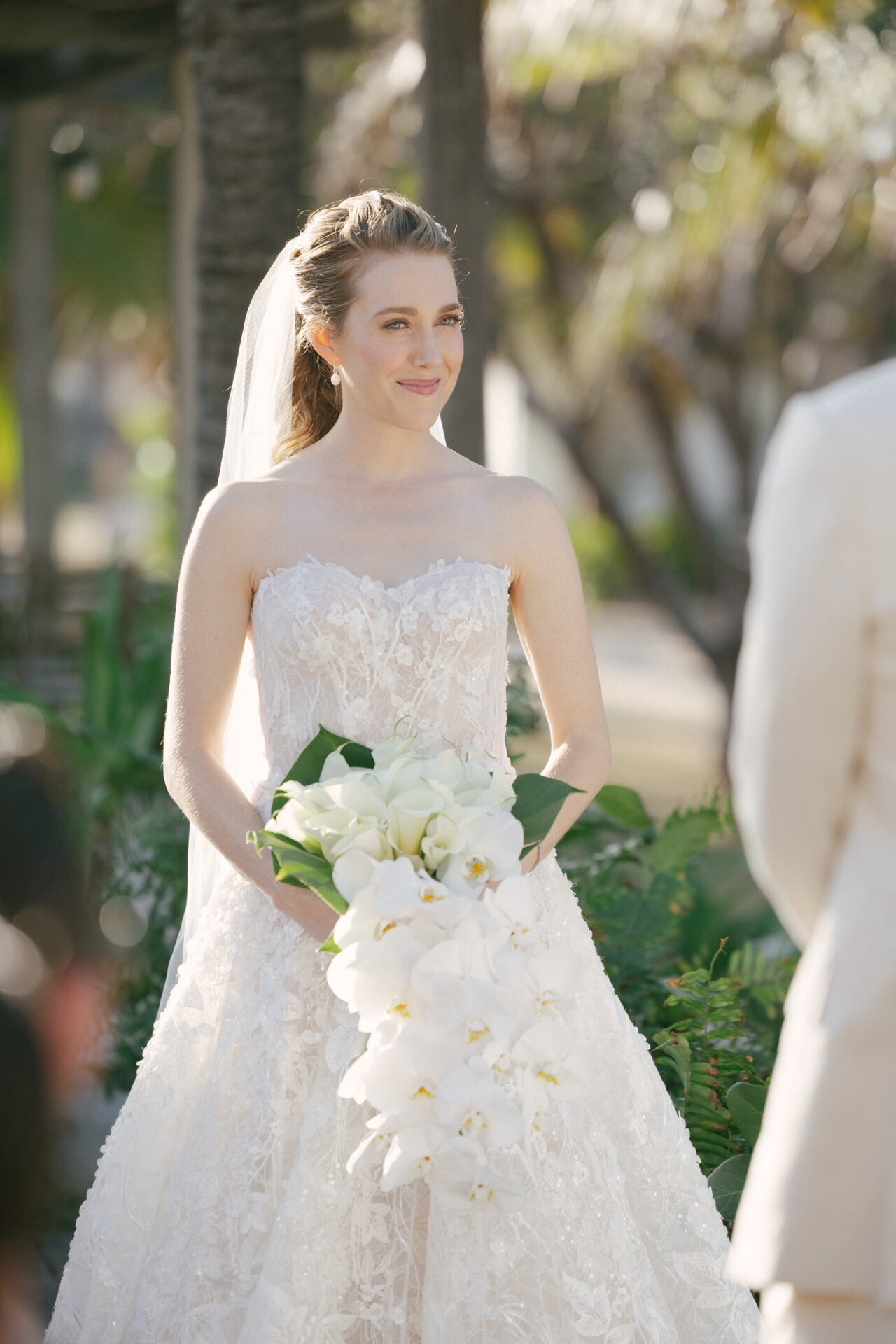 Bride in strapless lace dress holds white bouquet outdoors.