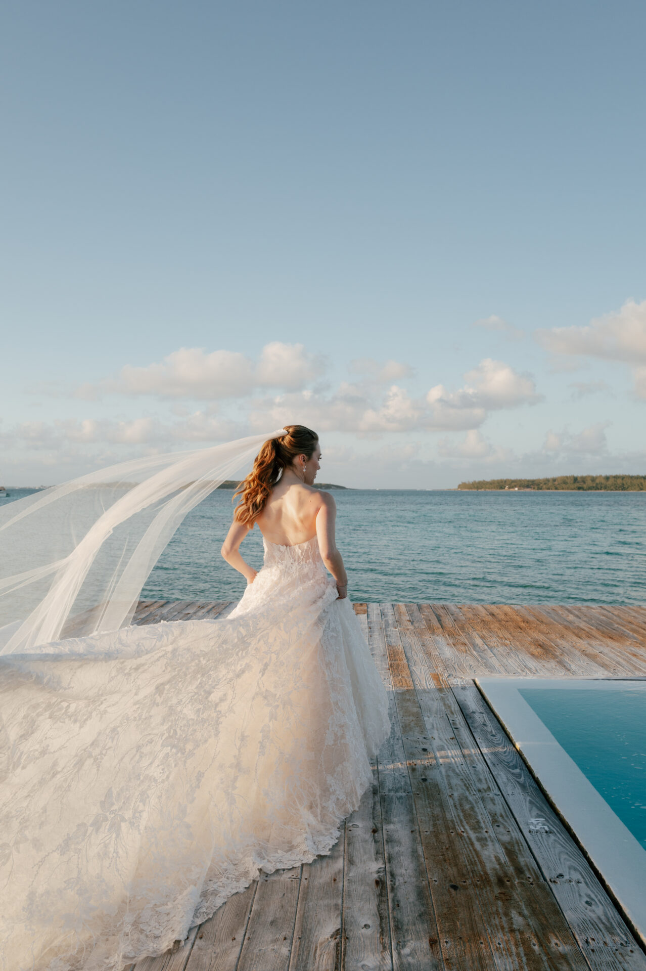 Bride in lace gown and veil on dock by sea.