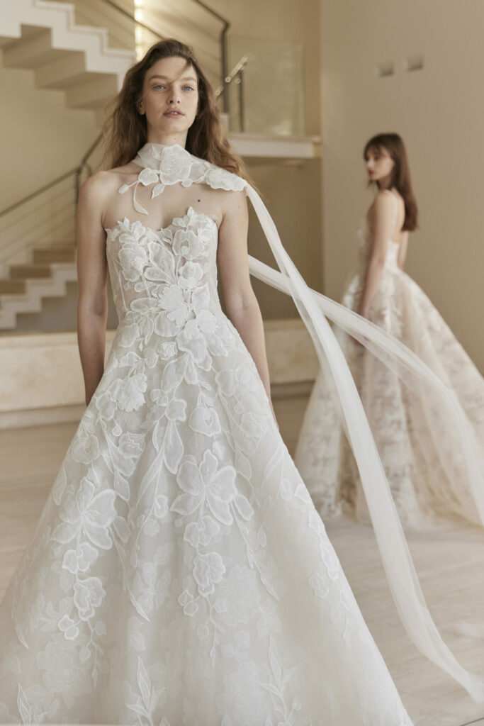 Two women model white, strapless lace gowns indoors.