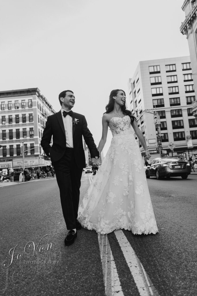 Bride and groom cross city street in wedding attire.
