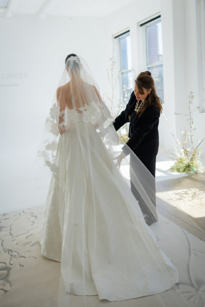 Bride in floral veil, sunlit room, another adjusts.