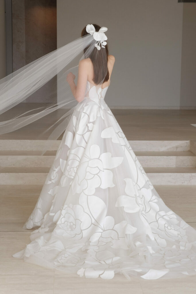 Bride in floral white dress with veil, facing away.