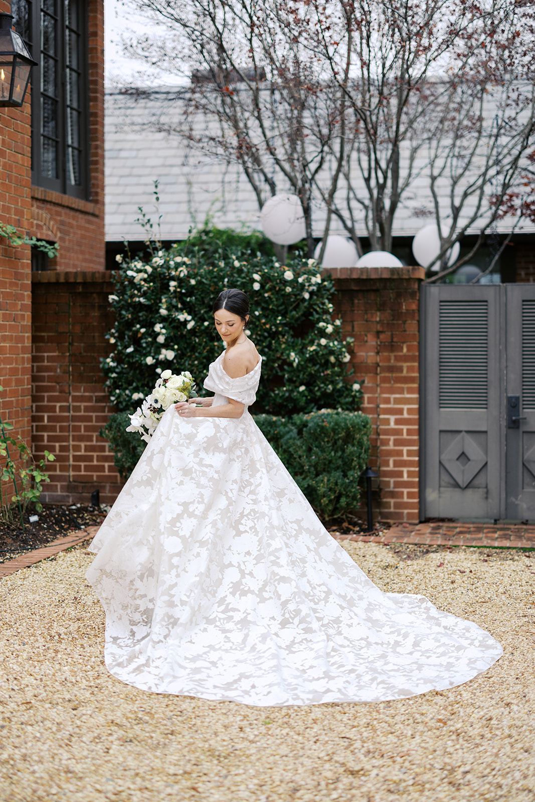 Bride in white floral gown smiles in courtyard.