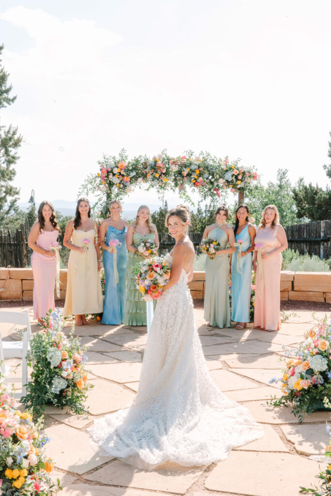 Bride poses with bouquet; bridesmaids under floral arch.
