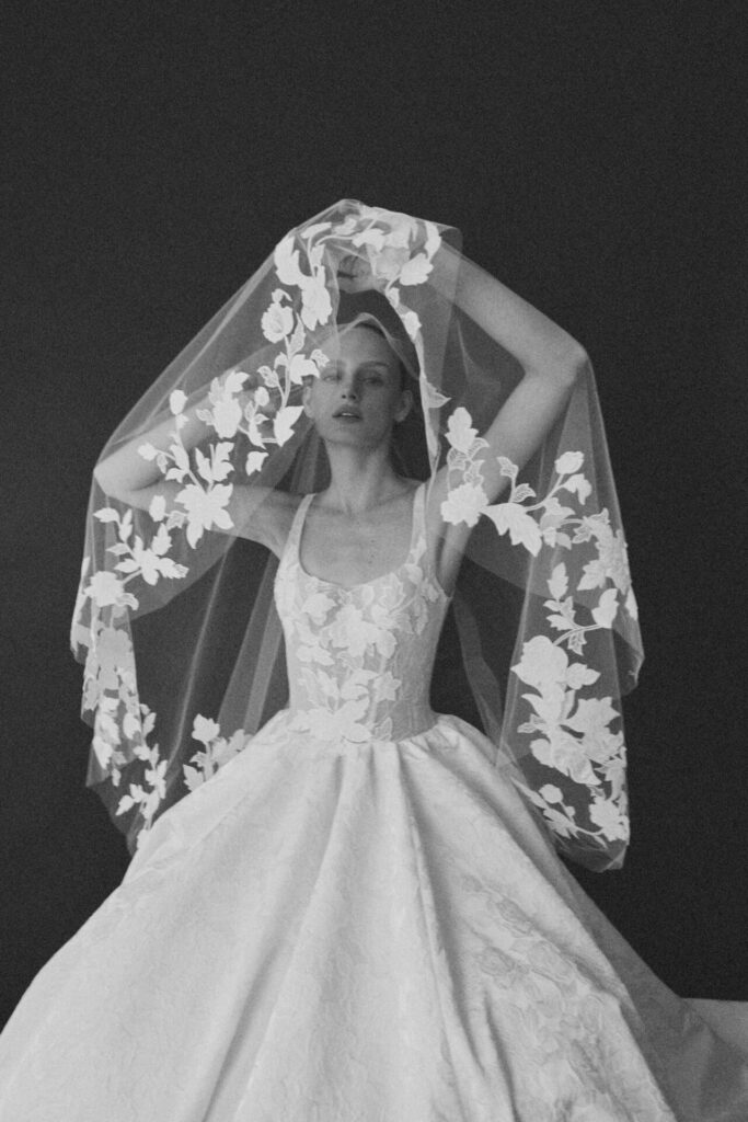 Woman lifts sheer veil in floral wedding gown, black-and-white.