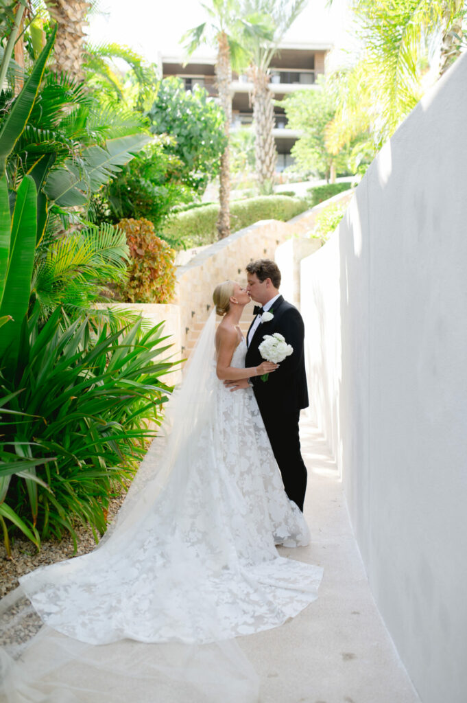 Bride and groom kiss outdoors in sunlight, greenery surrounds.